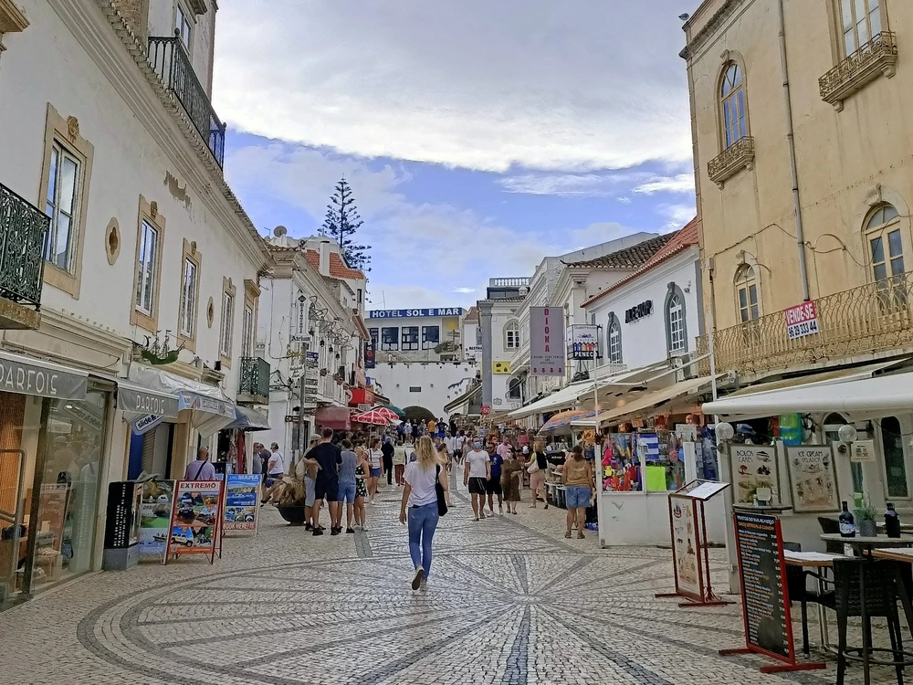 Tourists strolling down a charming cobblestone street in the Old Town of Albufeira, lined with shops and traditional Portuguese buildings.
