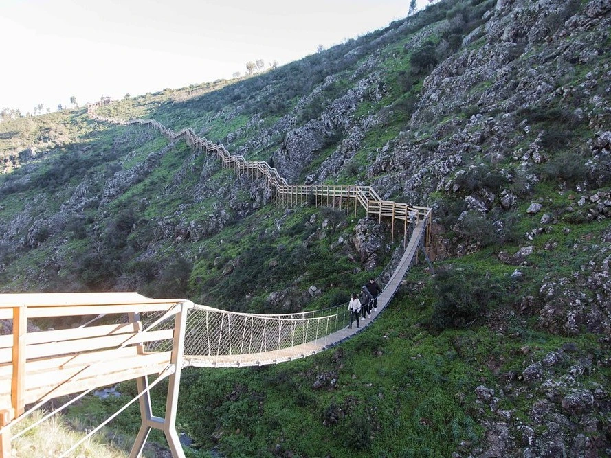 Hikers crossing the suspension bridge at the Barranco do Demo Boardwalk on the green, rocky hillsides of Monchique.