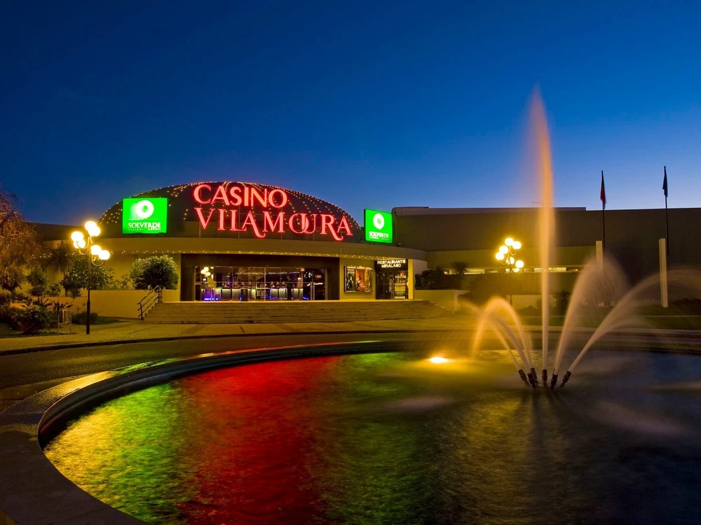 The vibrant entrance to Casino Vilamoura at night, illuminated by its large red neon sign and a colorful fountain in the foreground.