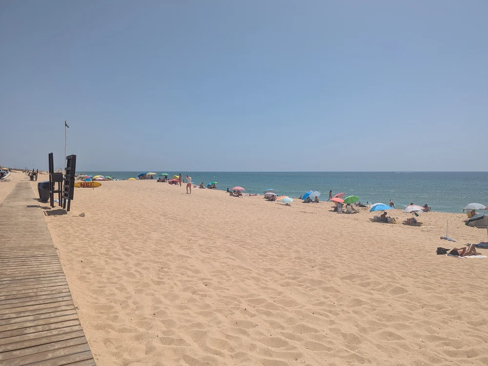 Sunbathers and colorful umbrellas spread across the golden sands of Praia de Faro (Faro Beach) on a clear summer day.