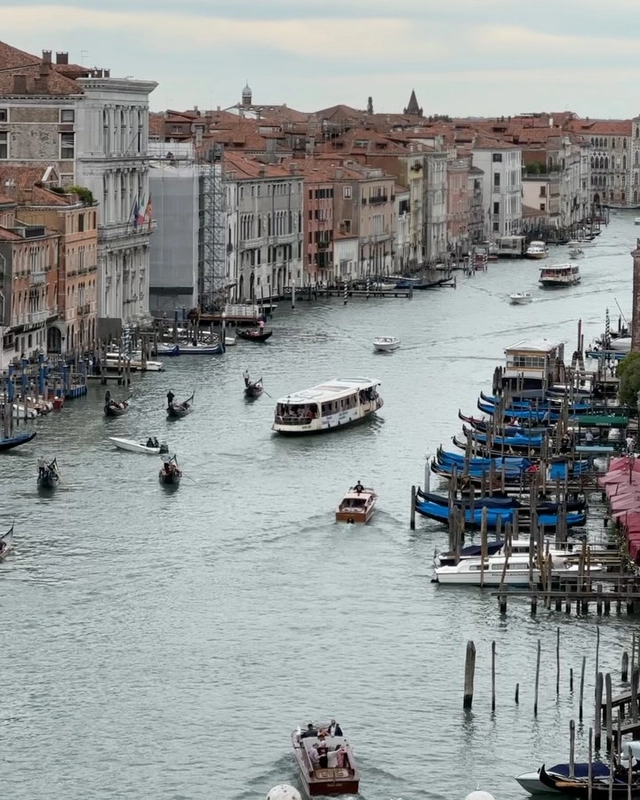 A panoramic rooftop view from Fondaco dei Tedeschi overlooking the Grand Canal in Venice, showing vaporetto boats, gondolas, and historic buildings lining the waterway.