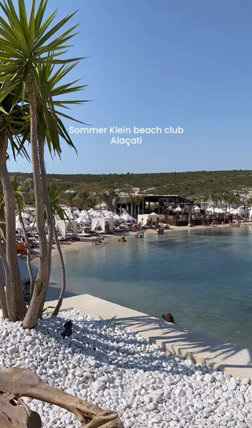 A scenic view of Sommer Klein beach club in Alaçatı, Turkey, with white cabanas lining the calm, clear water of the Aegean coast.