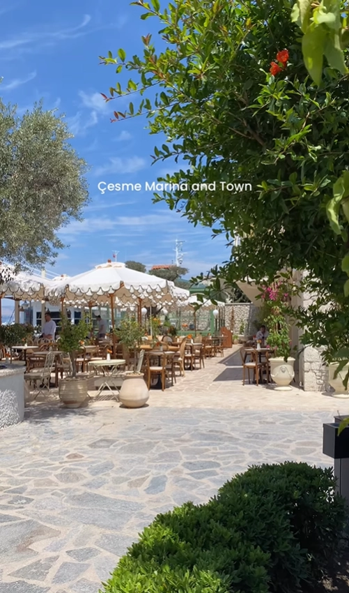 A charming outdoor cafe at the Çeşme Marina in Turkey, with elegant seating on a stone terrace surrounded by greenery.