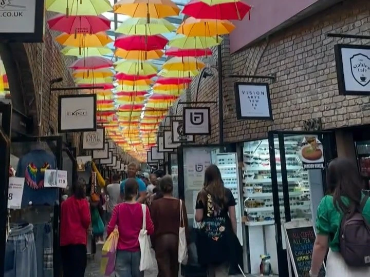 Shoppers exploring a vibrant alleyway in Camden Market, London, decorated with a ceiling of colorful red, orange, and yellow umbrellas.