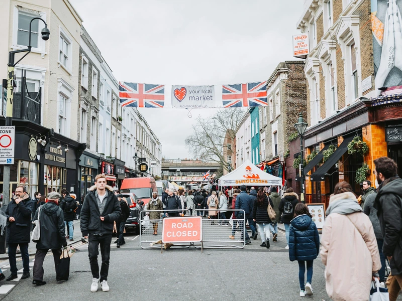 Crowds of shoppers walking down Portobello Road Market in London, with a Union Jack banner hanging overhead and market stalls lining the street.