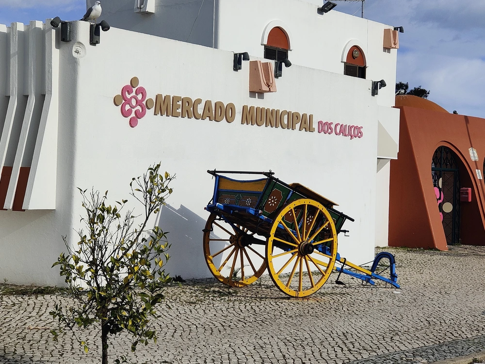 The white exterior of the Mercado Municipal dos Caliços in Albufeira, featuring a traditional, colorful wooden cart on the cobblestone plaza.