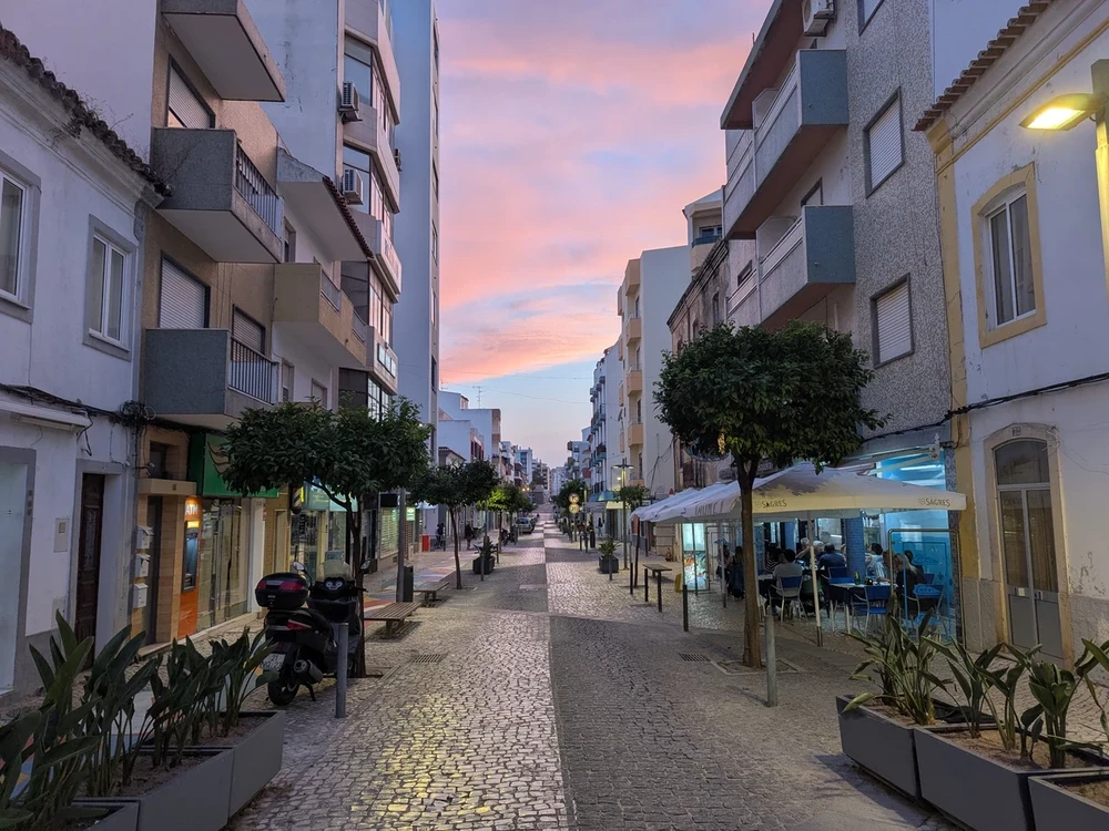 Charming cobbled street in downtown Portimão during sunset, with locals dining at outdoor cafés and soft pink skies overhead.