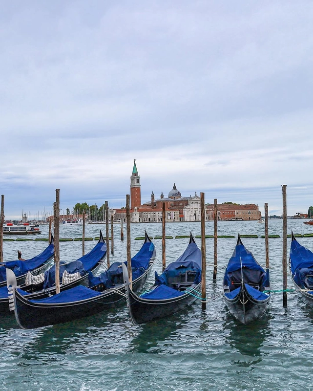 Gondolas with blue covers are moored along the Riva degli Schiavoni in Venice, with the island and church of San Giorgio Maggiore visible in the distance across the water.