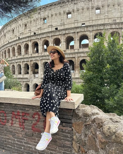 Beantowntraveller, in a floral dress and straw hat, smiles while sitting on a brick wall with the iconic Colosseum in the background, showcasing a blend of famous sites and non-touristy things in Rome.