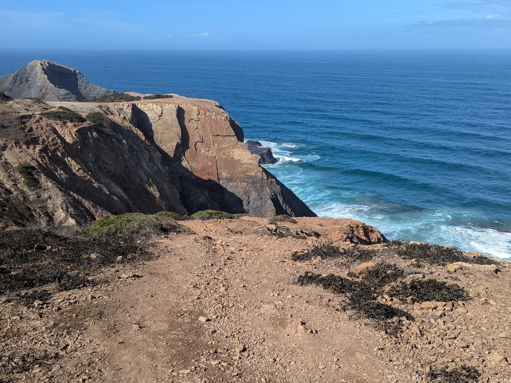 A dramatic view from the edge of a rugged, reddish-brown cliff on the Rota Vicentina Trail near Sagres