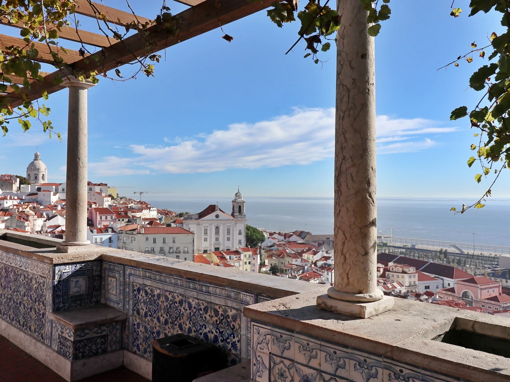 A scenic view from the Miradouro de Santa Luzia lookout point in Lisbon, overlooking the red-roofed houses of the Alfama district and the Tagus River.