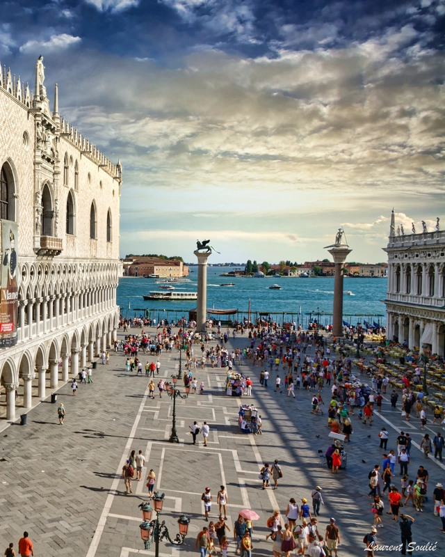 A wide view of St. Mark's Square in Venice, bustling with tourists, showing the Doge's Palace on the left and the waterfront in the background under a dramatic sky.