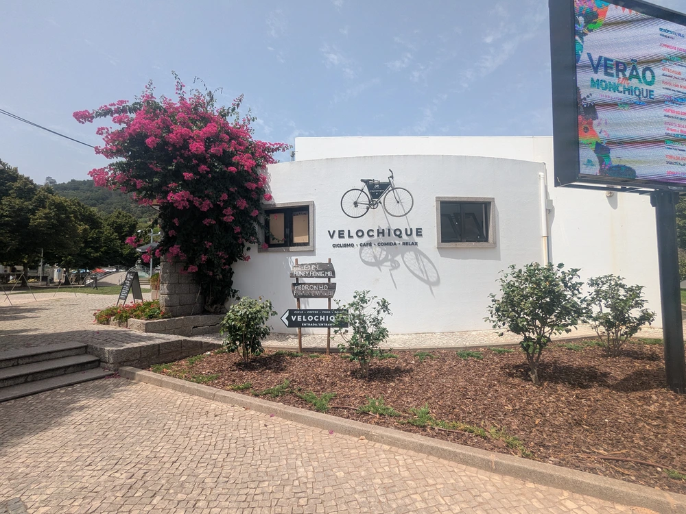 The modern white exterior of the Velochique cafe in Monchique, with its bicycle logo and a bright pink bougainvillea.